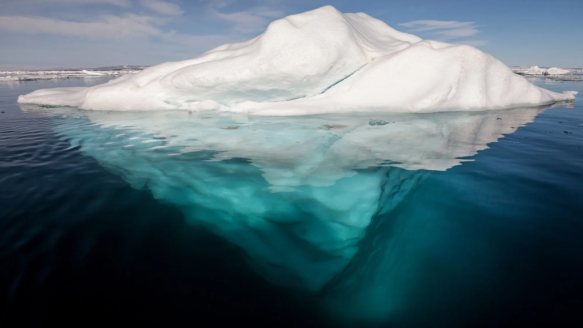 An iceberg photographed from the side: a bright tip above the waterline and a much larger mass below it fading into deep blue water