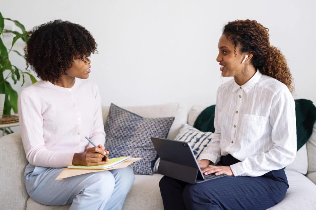 The ripple effect of coaching underrepresented women. Image is two Black women in business attire turned toward each other during a coaching session